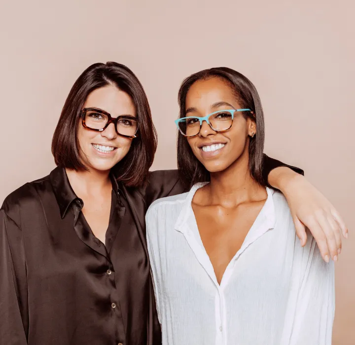 Two smiling women wearing glasses, one with a black satin shirt and the other with a white shirt, embracing each other.