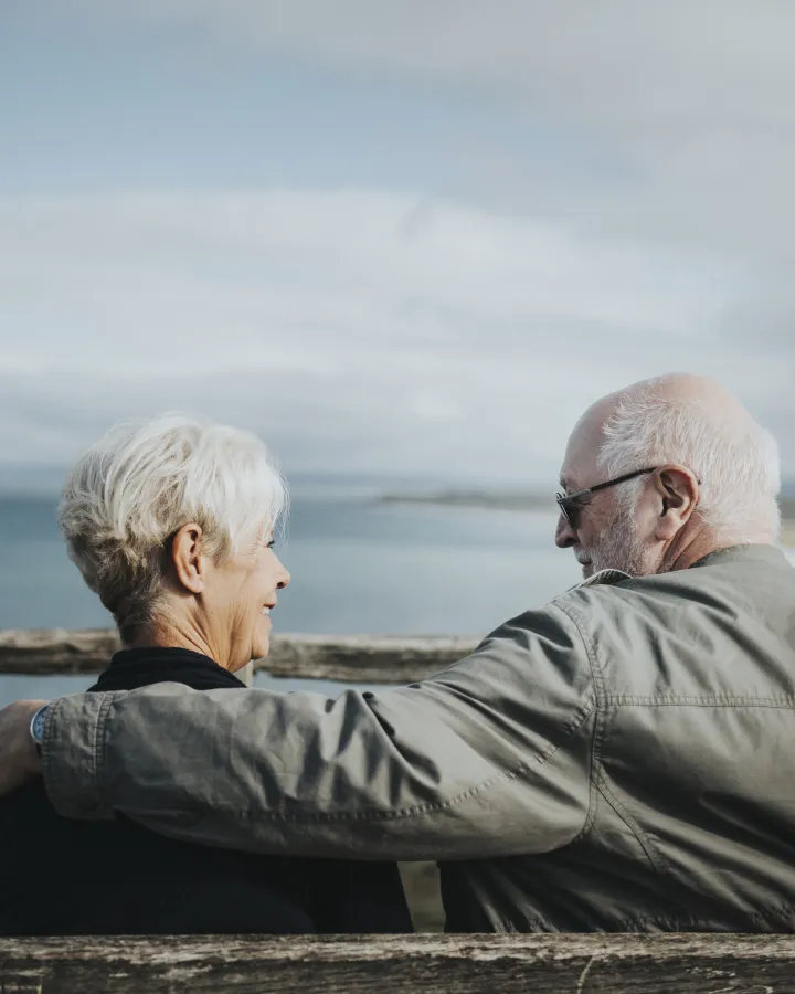 Elderly couple sitting on a bench facing a body of water with the man’s arm around the woman.