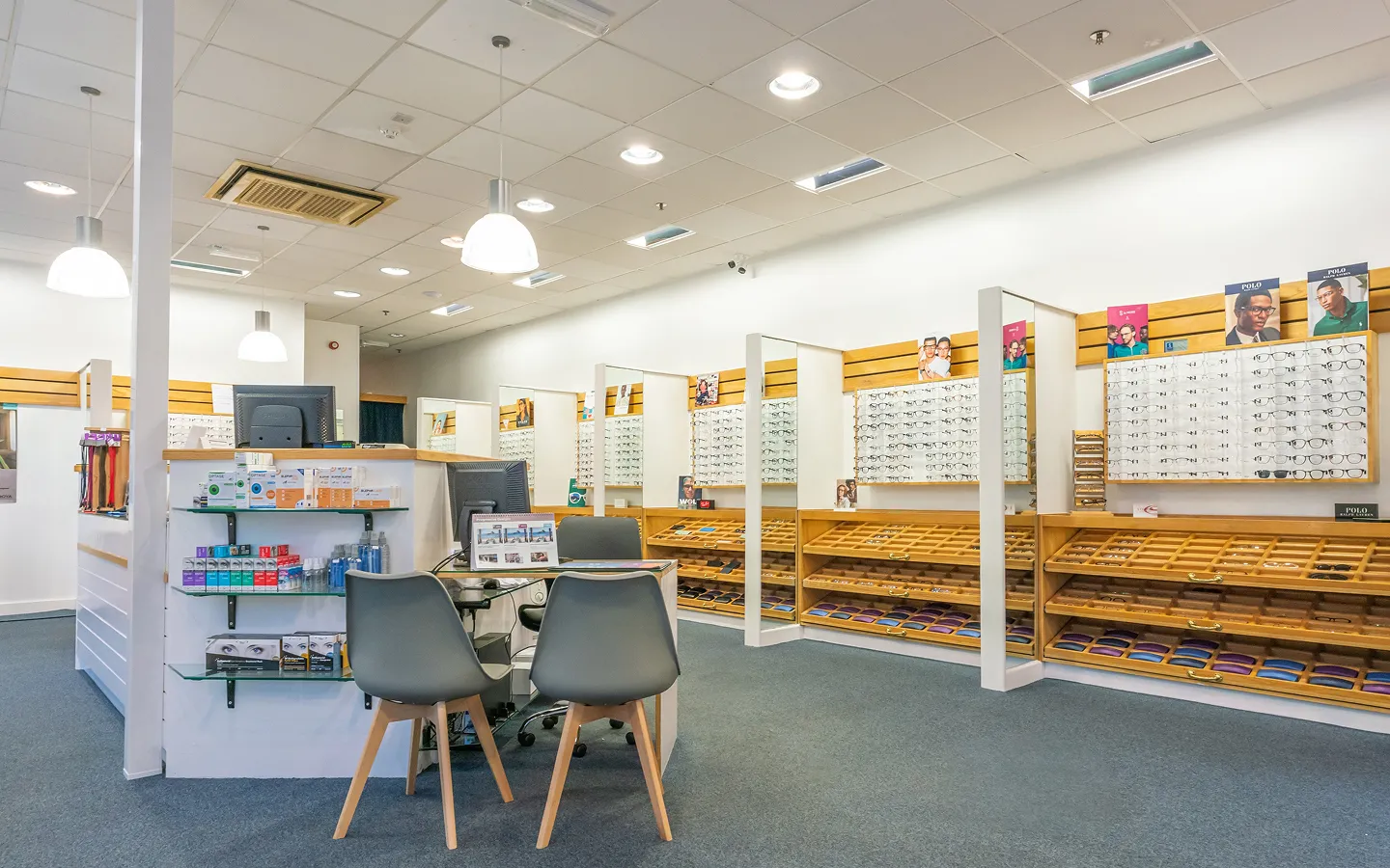 Bright optician store interior with display racks full of eyeglass frames and a consultation desk with two gray chairs.
