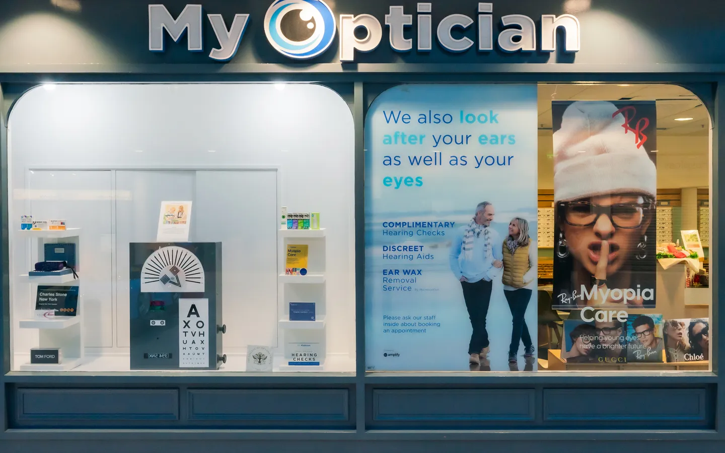Storefront of My Optician with eye and hearing care advertisements featuring a couple and vision test charts in the window.