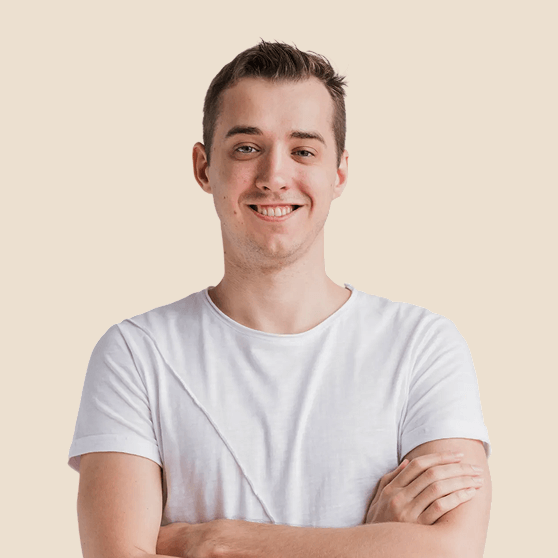 Young man with short hair smiling and crossing his arms, wearing a white t-shirt against a beige background.