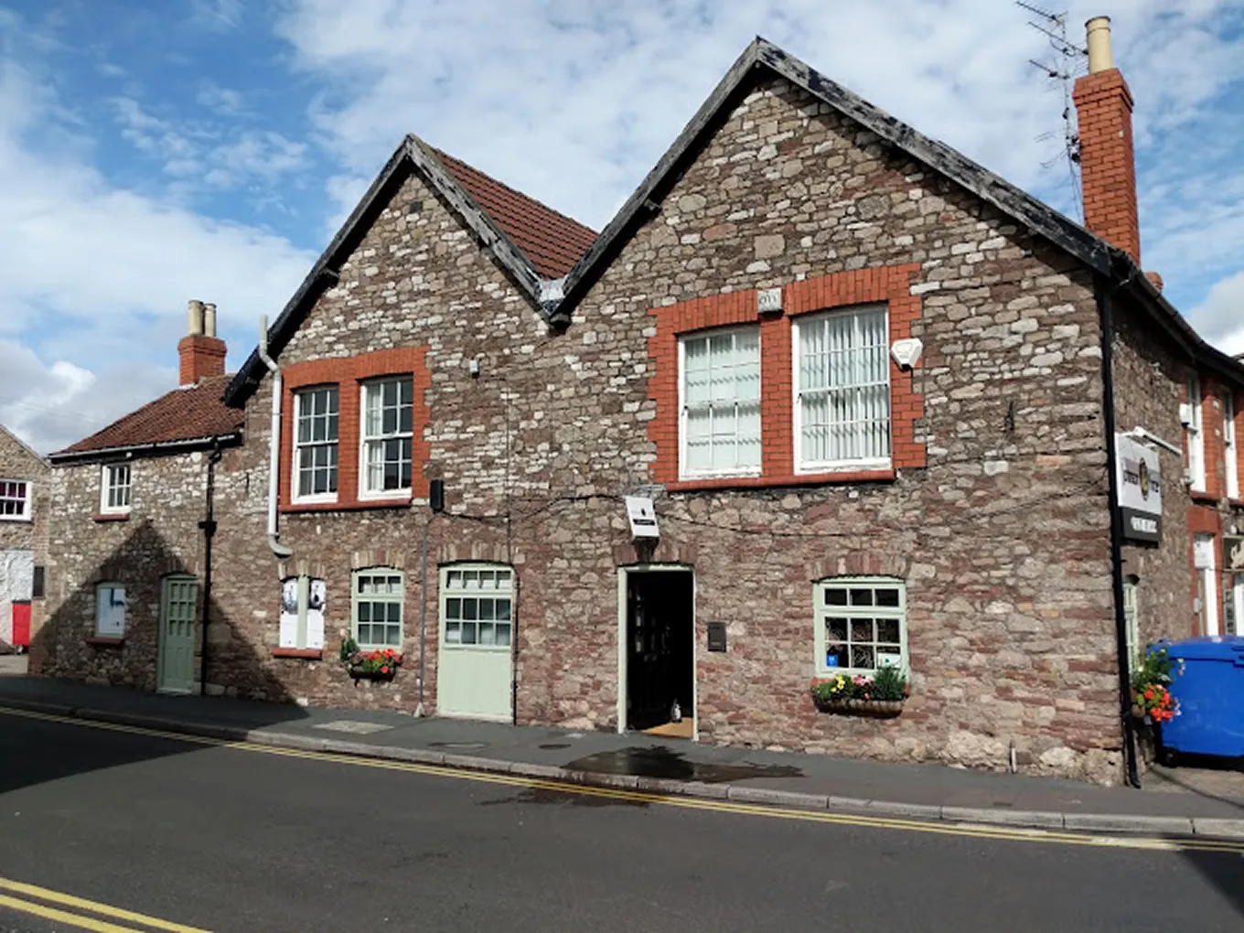 Stone building, red brick window frames, green doors, flower boxes, blue recycling bin, partly cloudy sky.