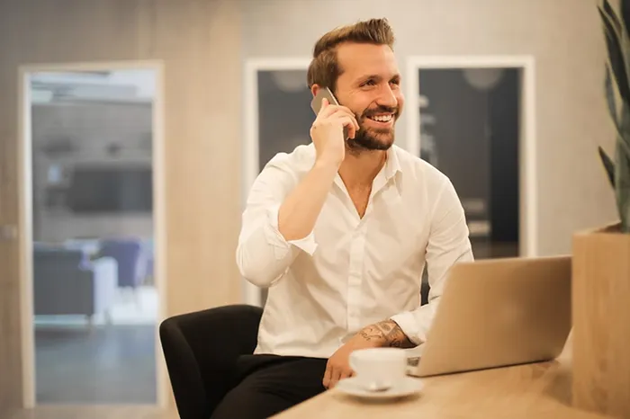 Smiling man in white shirt talking on a smartphone while sitting at a desk with a laptop and coffee cup.