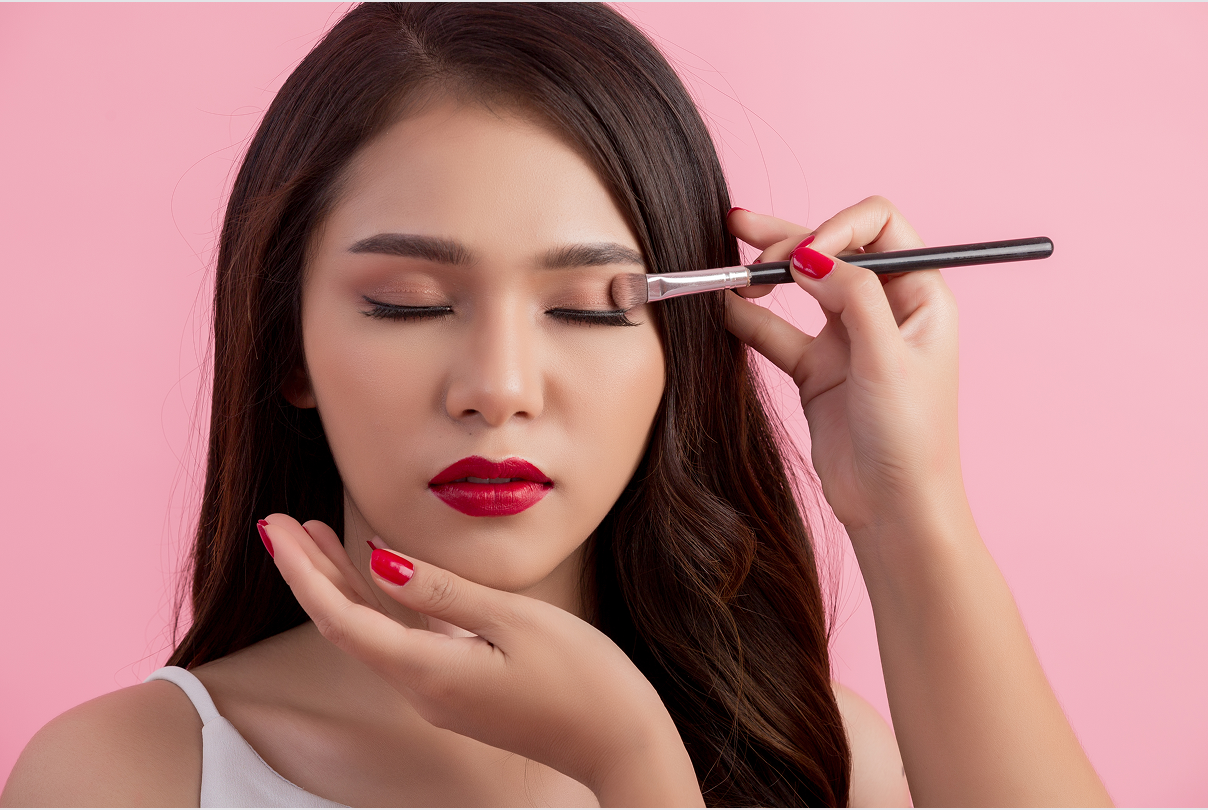 Woman with closed eyes having eyeshadow applied with a makeup brush on pink background.
