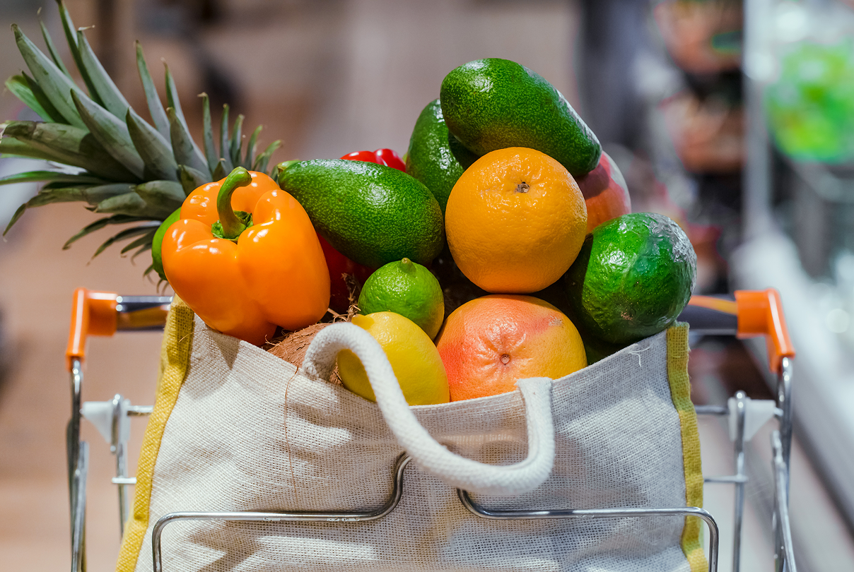 Reusable grocery bag filled with fresh fruits and vegetables including pineapple, bell peppers, avocados, lime, orange, lemon, and grapefruit inside a shopping cart.
