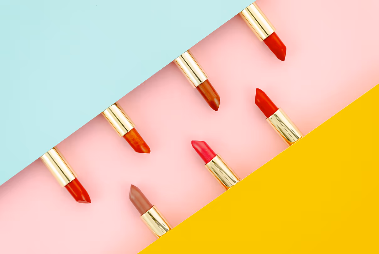 Six lipsticks with gold tubes in various shades of red and brown arranged diagonally on a pink background with blue and yellow paper edges.