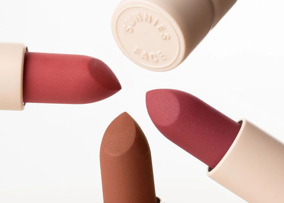Three lipsticks in shades of muted red, brown, and dark pink arranged around a beige lipstick cap embossed with 'SUNNIES FACE'.