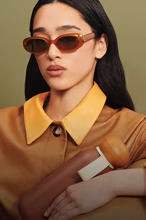 Woman with long dark hair wearing brown-tinted sunglasses and a brown coat with a yellow collar holding a matching brown and beige bag.