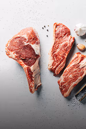 Three raw ribeye steaks on a light surface with garlic cloves and peppercorns scattered around.