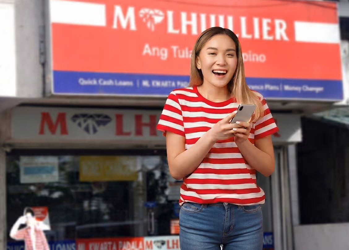 Smiling young woman in a red and white striped shirt using a smartphone standing in front of a MLhuillier storefront.