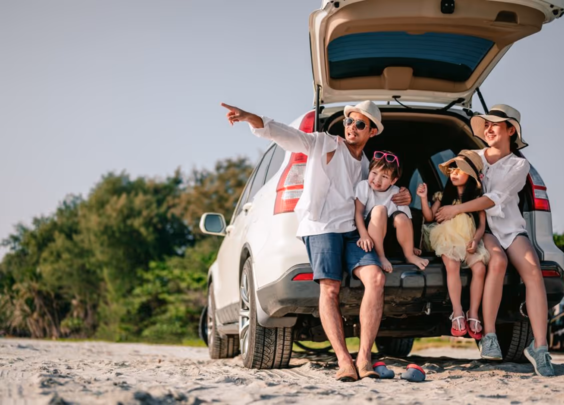 Family of four sitting in the open trunk of a white SUV on a sandy beach, with the father pointing towards the distance.