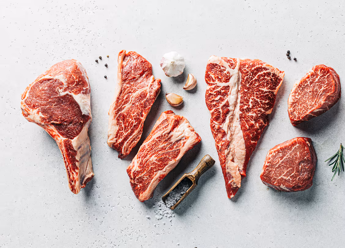Various raw marbled beef cuts arranged on a light surface with garlic cloves, black peppercorns, coarse salt in a wooden scoop, and rosemary sprigs.