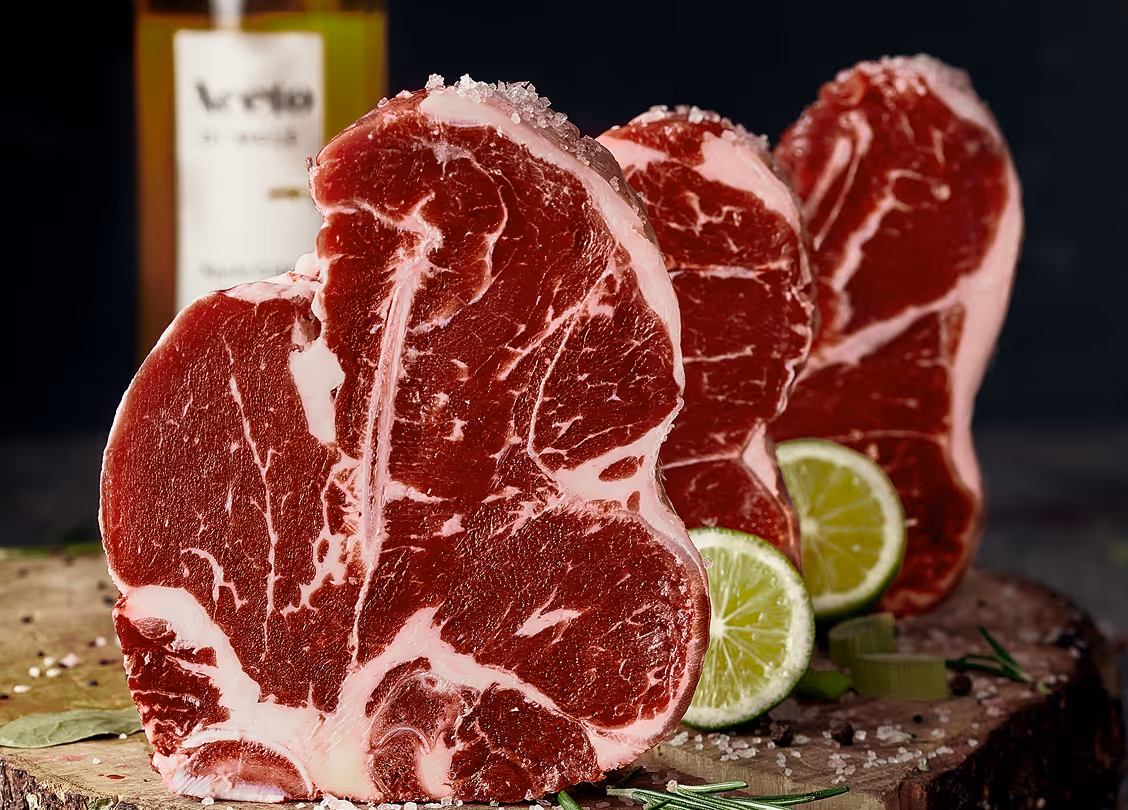Three raw ribeye steaks standing upright on a wooden cutting board with lime halves and rosemary sprigs.