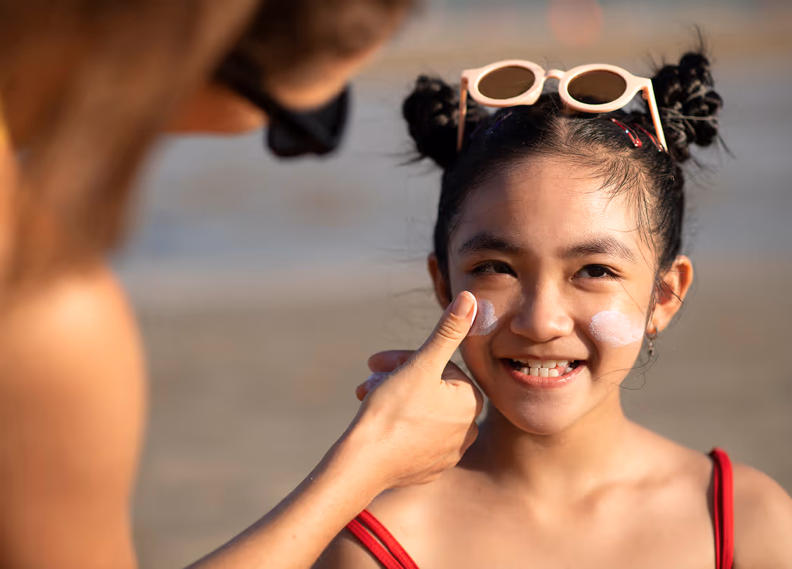 Young girl smiling on the beach with sunscreen being applied on her cheeks, wearing red straps and sunglasses on her head.