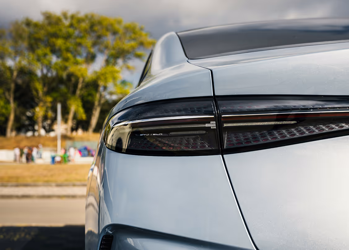 Close-up of the rear left side of a silver ByD electric vehicle showing its taillight and trunk with trees and people blurred in the background.