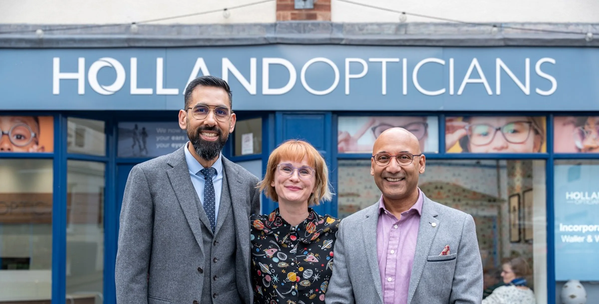 Three opticians wearing glasses smiling in front of Holland Opticians store with blue facade and signage.
