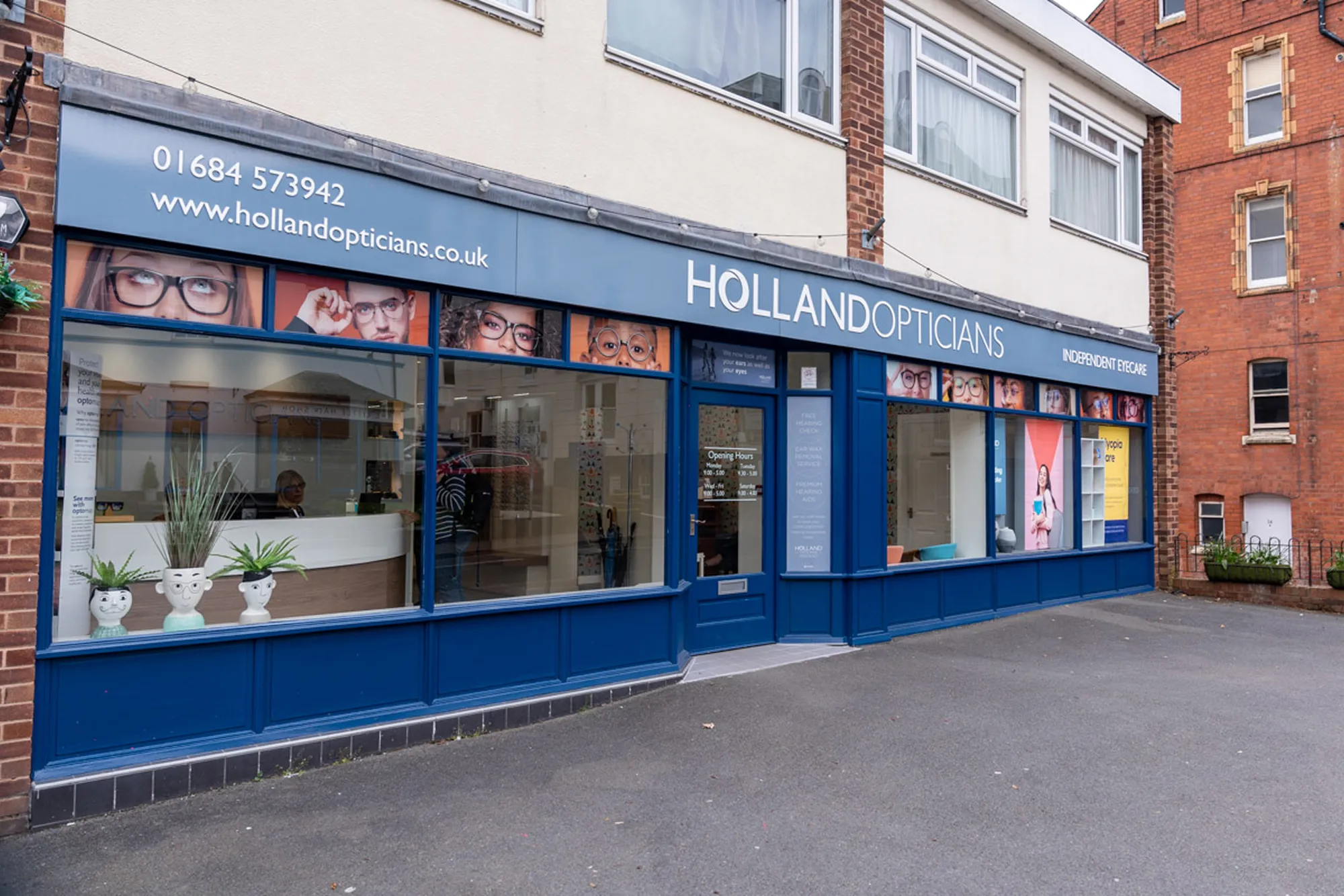 Holland Opticians storefront with blue facade, eyeglasses displays in windows, and face-shaped potted plants outside.