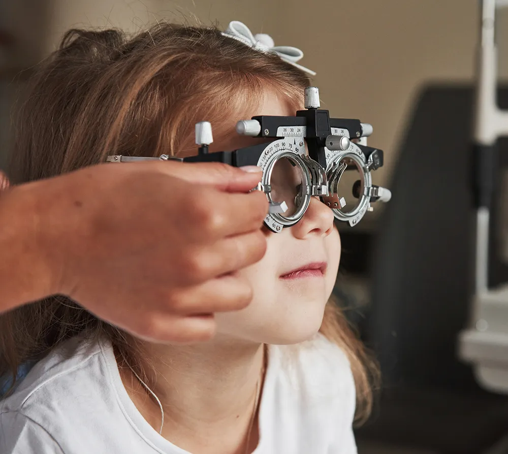 Child undergoing an eye exam with an optometrist adjusting a phoropter in front of her face.