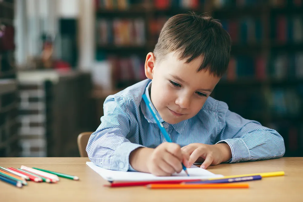 Young boy in a blue striped shirt drawing with colored pencils at a table indoors.