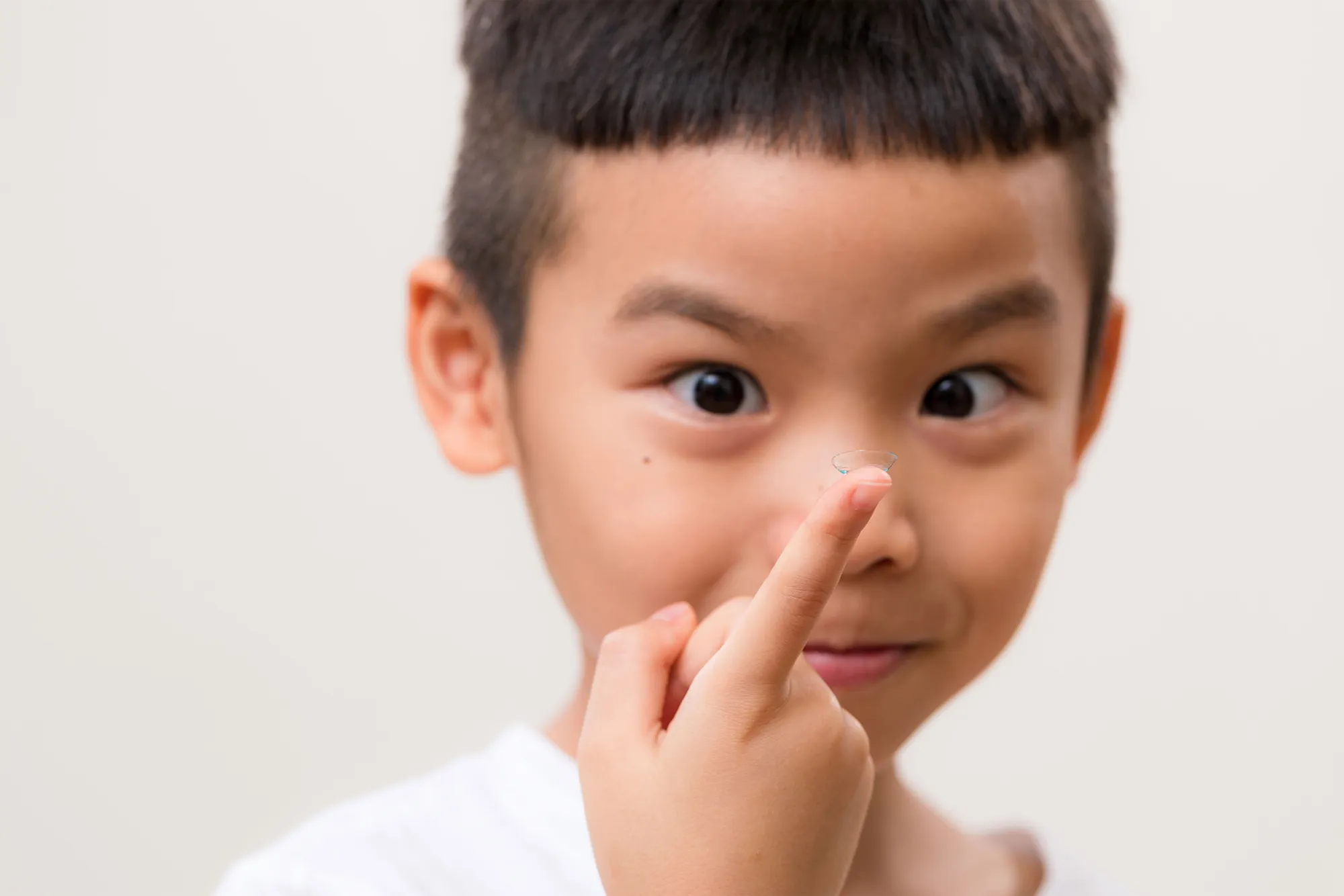 Young boy holding a clear contact lens on his fingertip, focusing on the lens with a playful expression.