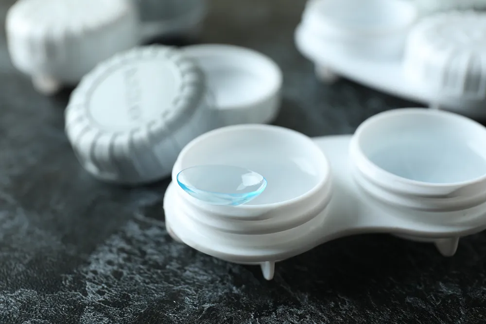 Close-up of a clear contact lens resting on a white plastic contact lens case on a dark textured surface.