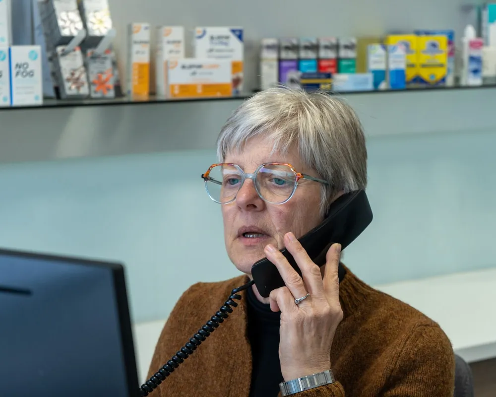 Older woman with gray hair and glasses talking on a corded phone, with product boxes on a shelf behind her.