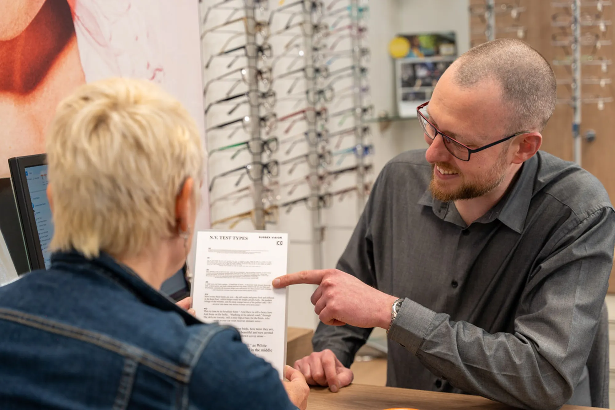 Man in glasses pointing at a paper held by a woman in an eyewear store with glasses displayed in the background.