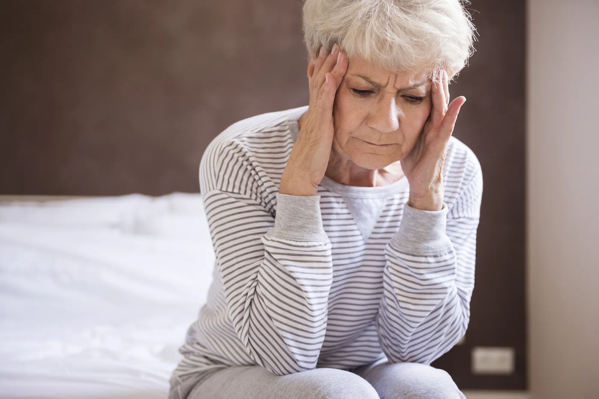 Elderly woman sitting on a bed, holding her temples with a pained expression, suggesting a headache or stress.