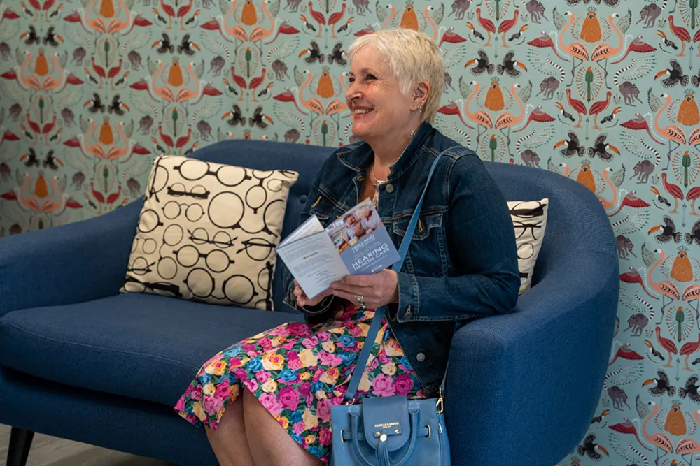 Smiling woman in a floral dress and denim jacket sitting on a blue couch, holding a healthcare brochure.