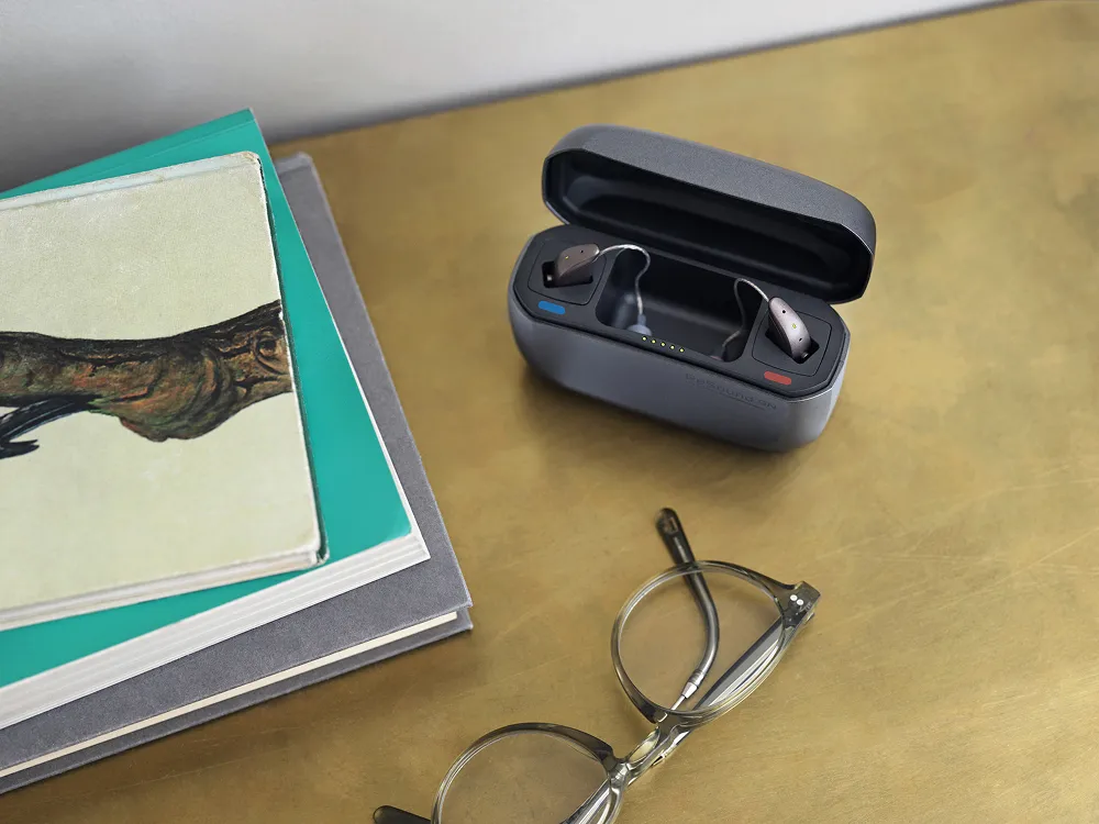 Charging case with two behind-the-ear hearing aids on a wooden surface next to a pair of eyeglasses and a stack of books.
