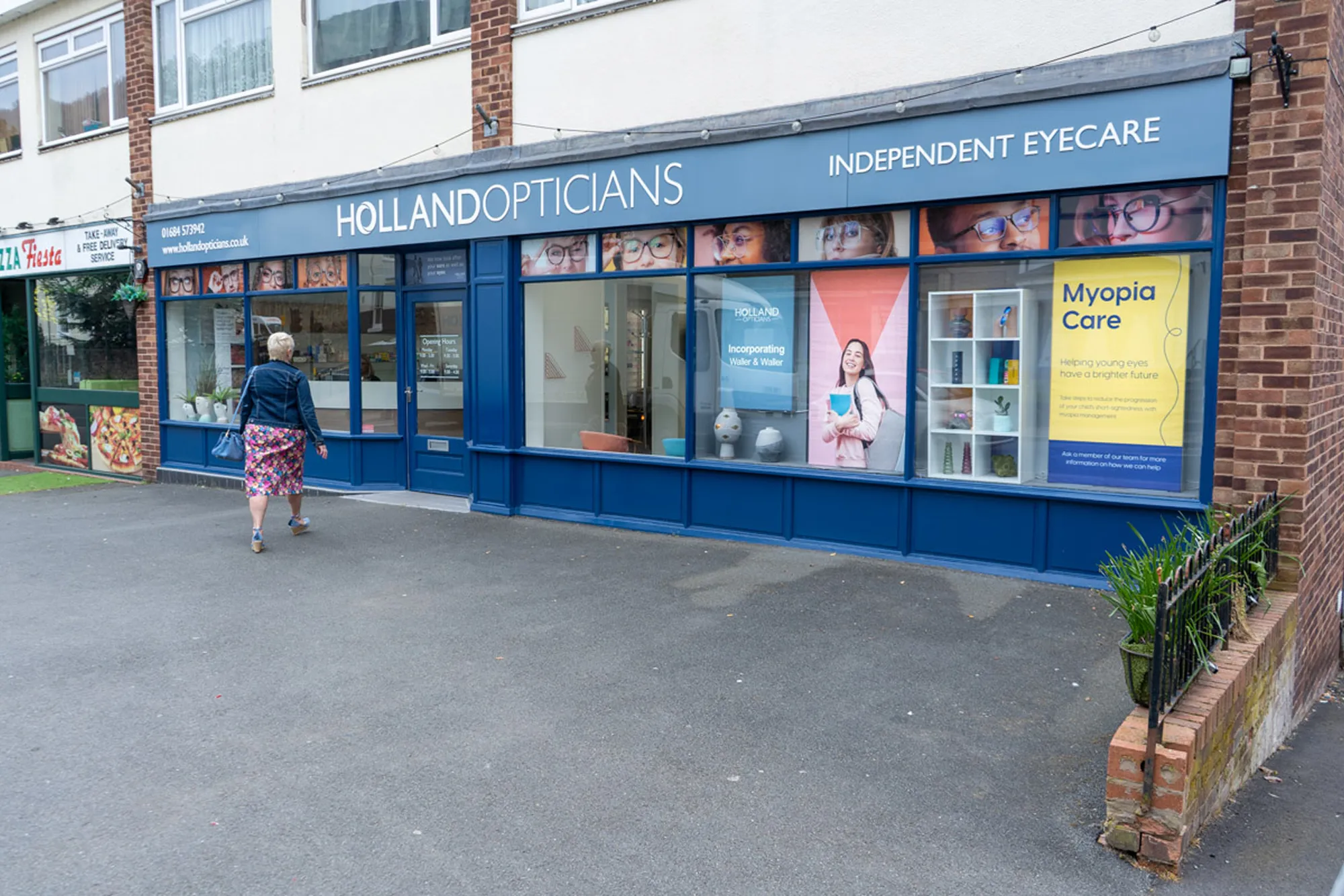 Exterior view of Holland Opticians with blue storefront, window displays featuring glasses and advertisements, and a person walking towards the entrance.