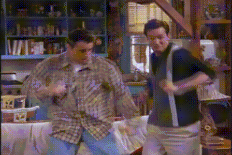 Two men energetically dancing in a cozy living room with bookshelves and a window in the background.