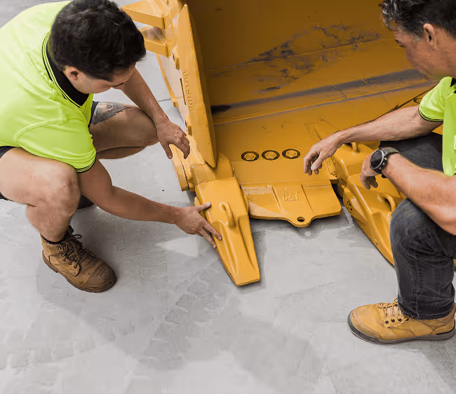 Two men in neon yellow shirts inspecting the yellow metal teeth of heavy machinery on a concrete floor.