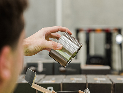 Person holding a silver metal cup above an open black product box on a table filled with similar boxes.