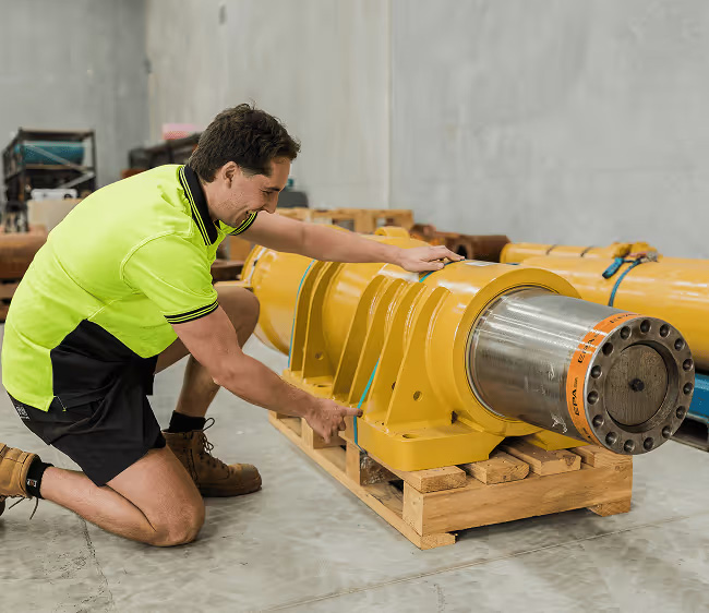 Man in high-visibility shirt kneeling and measuring a large industrial yellow mechanical component on a wooden pallet labeled Gateway Group WA.