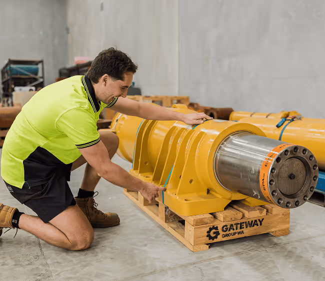 Man in high-visibility shirt kneeling and measuring a large industrial yellow mechanical component on a wooden pallet labeled Gateway Group WA.