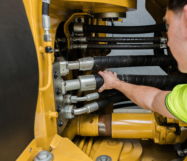 Person wearing a neon green shirt working on hydraulic hoses of yellow heavy machinery.