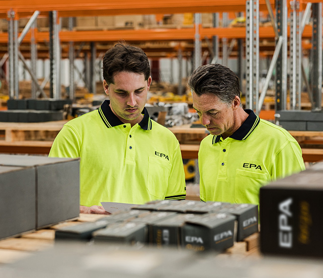 Two men in yellow EPA polo shirts examining documents in a warehouse with shelves and boxes.