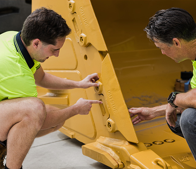 Two men in yellow shirts inspecting and pointing at a large yellow industrial machine component.