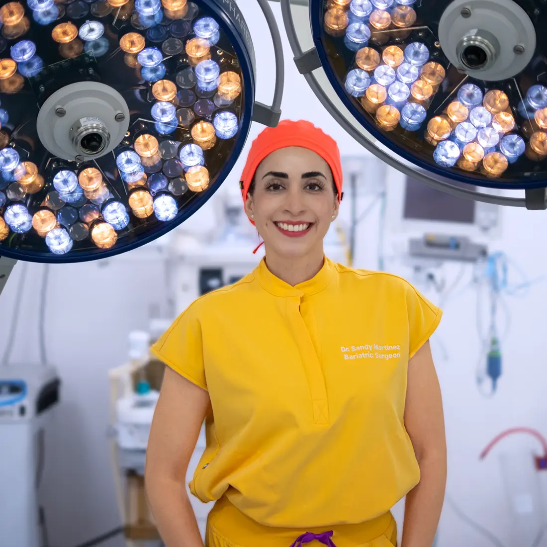 Smiling female surgeon in yellow scrubs and red surgical cap standing in operating room under surgical lights.