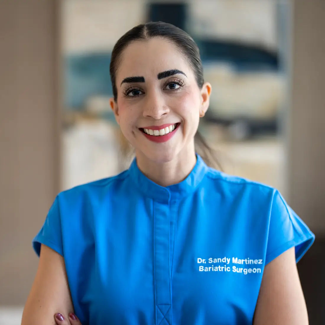 Smiling woman with dark hair pulled back wearing a blue medical uniform with 'Dr. Sandy Martinez Bariatric Surgeon' embroidered.