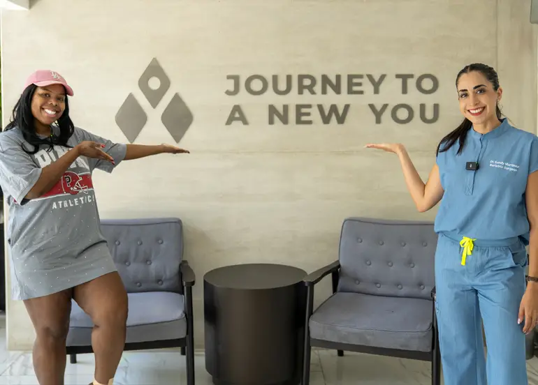 Two women smiling and gesturing towards the "Journey to a New You" sign at Dr. Sandy Bariatrics, celebrating the life-changing results of personalized weight loss surgery in Tijuana.