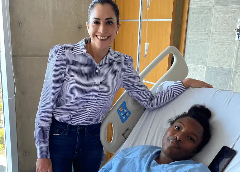 Smiling woman standing beside a hospital bed where a patient in blue hospital gown is lying down.