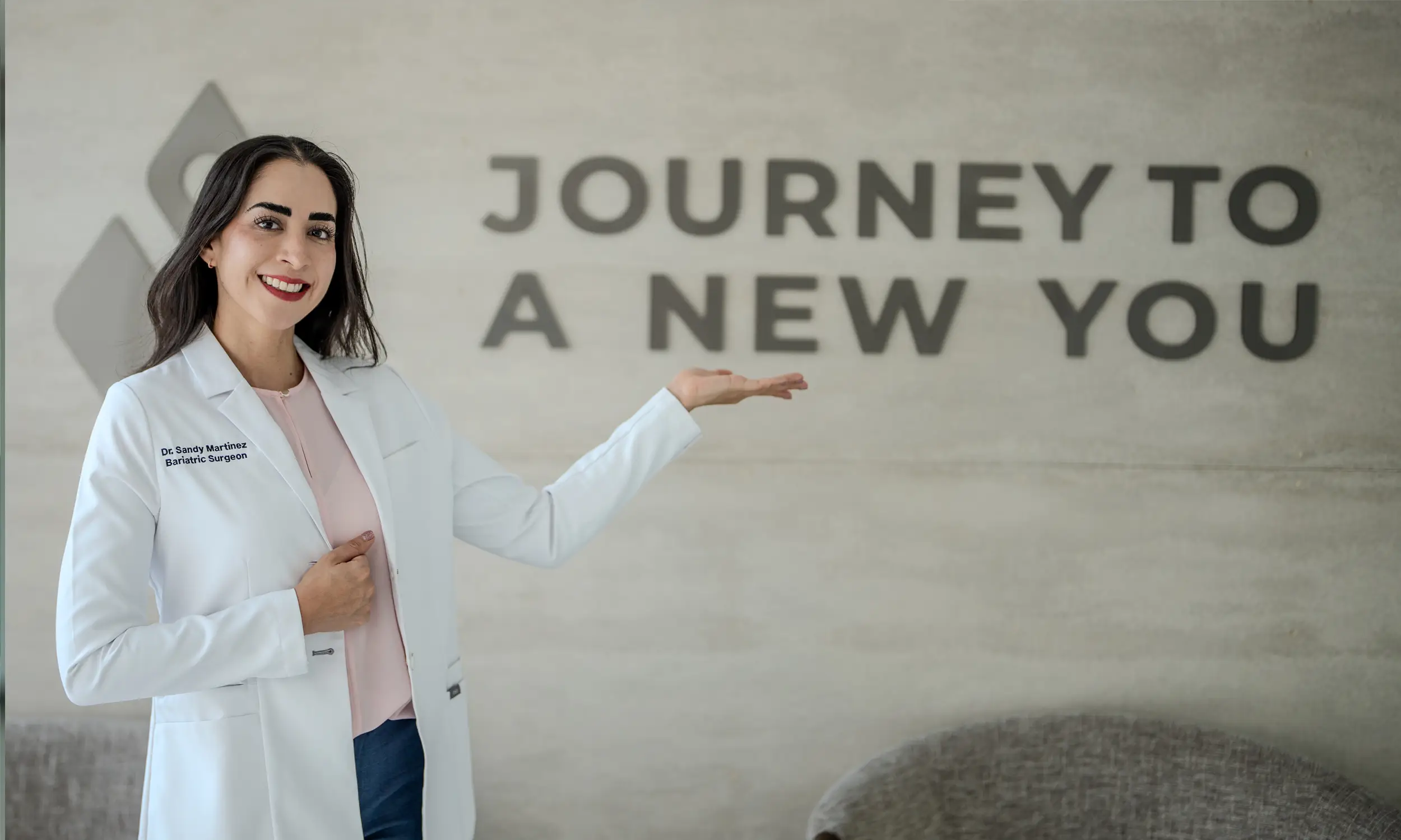 Smiling female bariatric surgeon Dr. Sandy Martinez in a white coat gesturing towards a wall sign that reads 'Journey to a New You'.