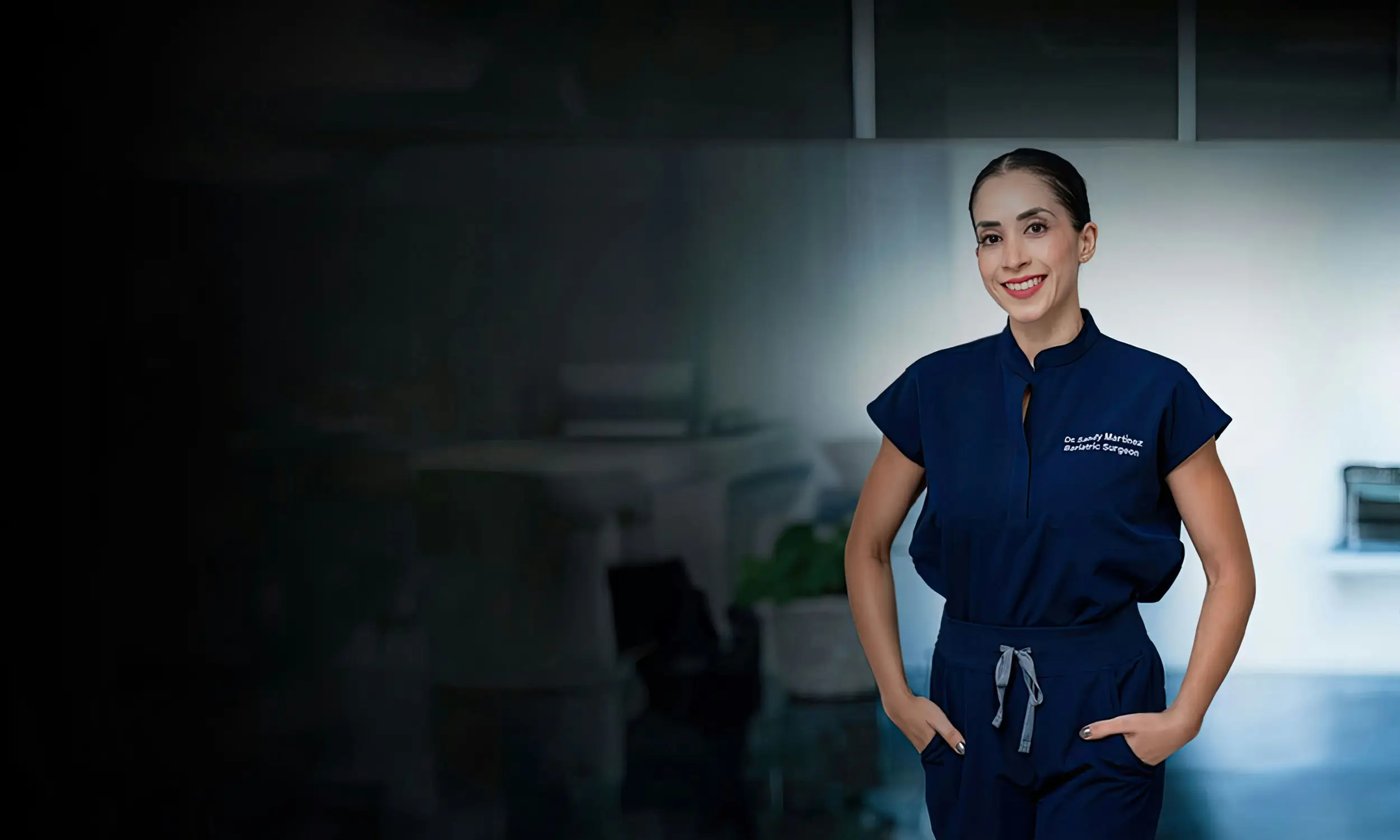 Smiling female doctor in navy blue scrubs with hands in pockets standing in a medical office.