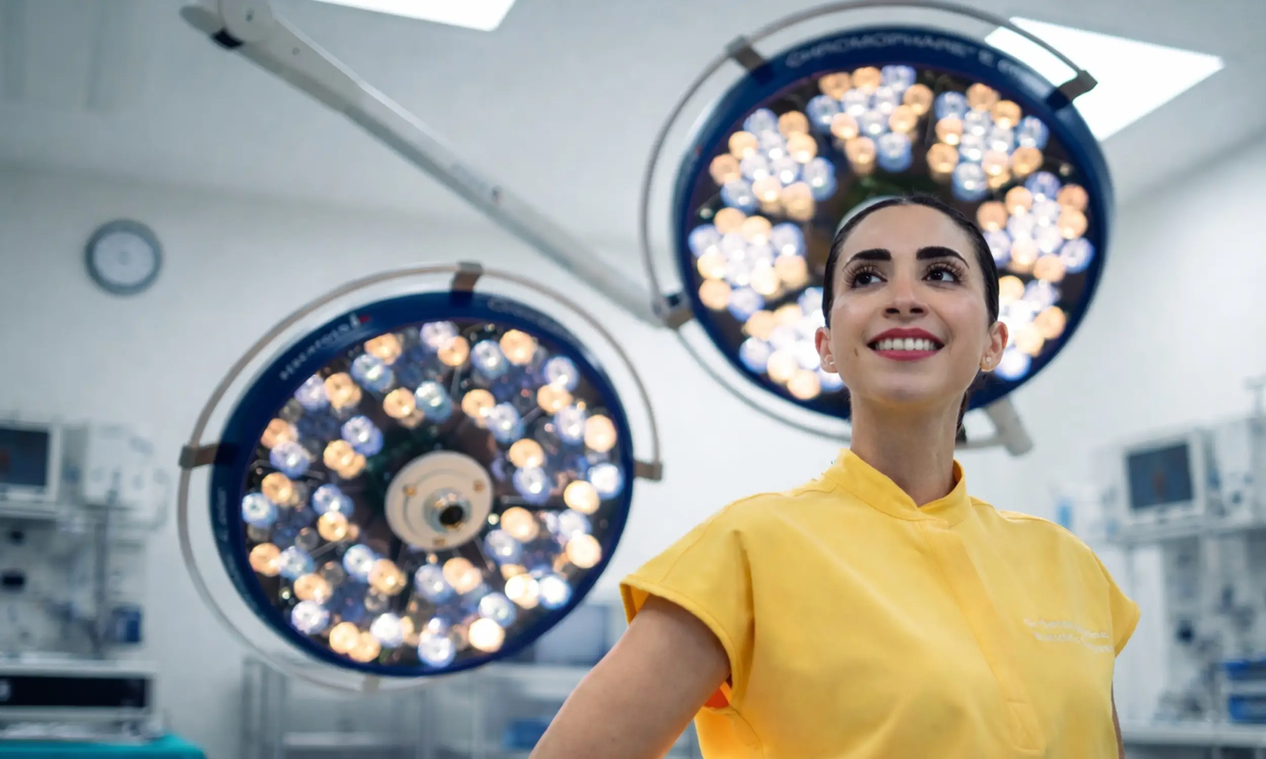 Smiling female medical professional in yellow scrubs standing in a brightly lit operating room with surgical lights overhead.