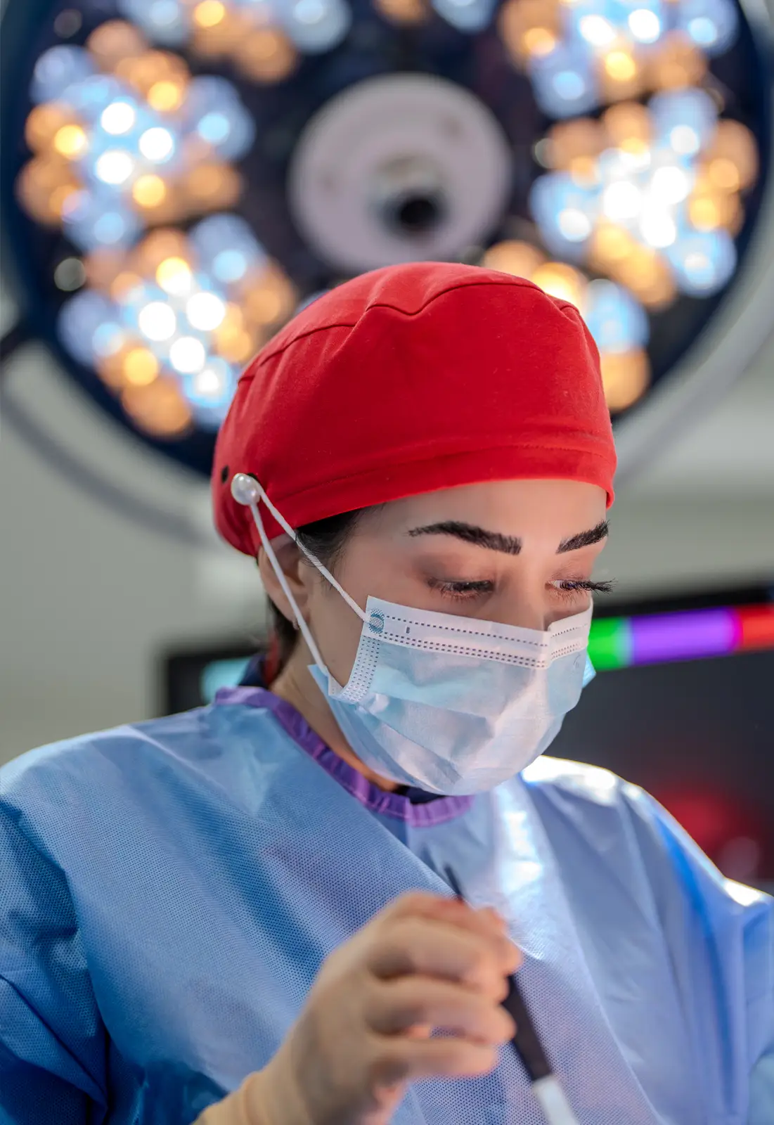 Female surgeon wearing a red surgical cap and blue gown, focused during an operation under bright surgical lights.