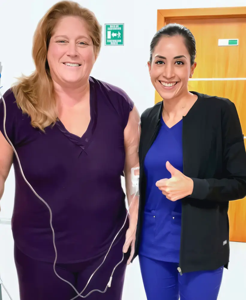 Smiling woman in purple outfit with IV line stands next to a smiling healthcare professional in blue scrubs giving a thumbs up.