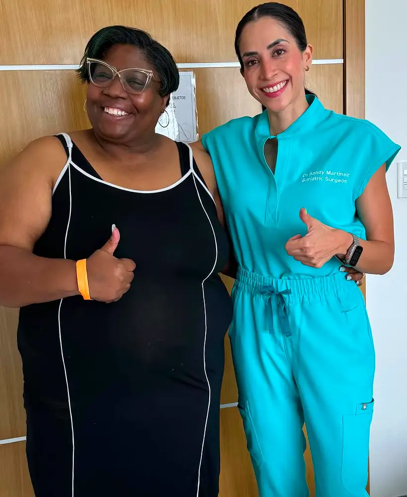Two smiling women standing next to each other and giving thumbs up, one wearing a black dress and the other in turquoise medical scrubs.