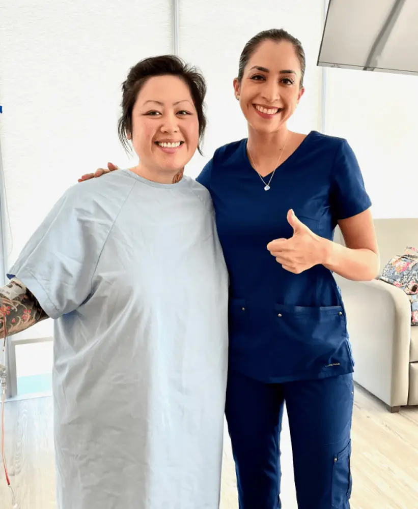 Two smiling women standing together, one in a light hospital gown and the other in blue medical scrubs giving a thumbs up.